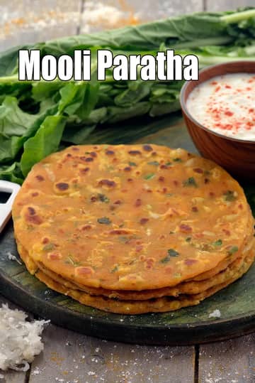 A stack of three golden-brown, pan-fried Mooli Parathas (radish-stuffed flatbreads) is displayed on a rustic dark wooden board. The parathas show visible bits of green herbs and radish throughout the dough. In the background, there is a bunch of fresh, leafy radish greens and a brown wooden bowl filled with creamy white curd topped with a sprinkle of red chili powder. To the left, a small pile of freshly grated white radish is visible on the wooden surface.