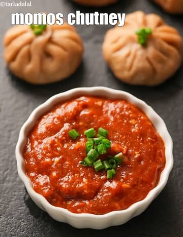 A white bowl filled with bright red momos chutney garnished with chopped spring onions, placed on a dark surface with steamed momos in the background.