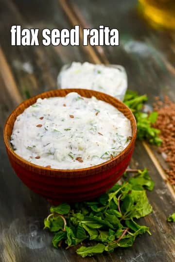 A wooden bowl filled with creamy Flax Seed Raita, garnished with roasted flax seeds and chopped herbs. The bowl sits on a rustic wooden surface surrounded by fresh green mint leaves and a small pile of flax seeds in the background.