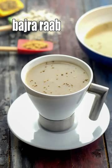 A white ceramic cup filled with Bajra Raab, a traditional Rajasthani and Gujarati warming drink made from pearl millet flour. The drink has a smooth, creamy texture and is garnished with carom seeds (ajwain). The cup is placed on a matching saucer over a dark wooden rustic table, with a red spatula and millet flour visible in the blurred background.
