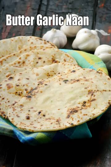 Butter garlic naan stacked on a cloth-lined plate, lightly charred and served with fresh whole garlic in the background.