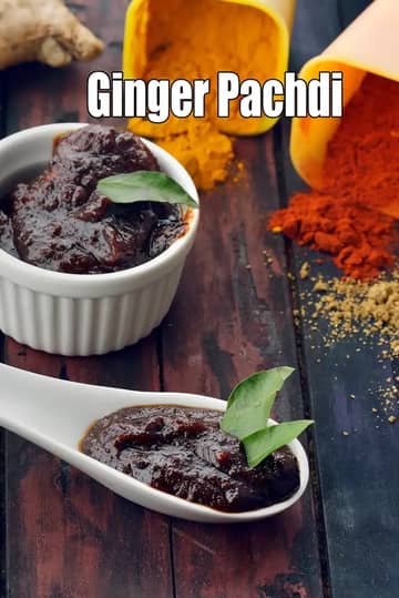 A close-up image of Ginger Pachdi (ginger chutney) served in a white ramekin and a ceramic spoon, garnished with fresh curry leaves. Fresh ginger roots are placed on the side, along with spilled spices like turmeric powder, red chilli powder, and coriander powder in small yellow containers on a rustic wooden background.