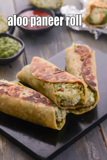 A close-up of three golden-brown Aloo Paneer Rolls stacked on a black serving board. The rolls are made with toasted flatbreads (rotis) filled with a visible stuffing of mashed potatoes, crumbled paneer, and fresh green herbs. In the background, small bowls of spicy green chutney and tangy red ketchup are served alongside, with another roll partially wrapped in aluminum foil.