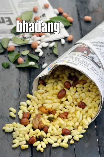 A close-up shot of Bhadang, a spicy puffed rice snack, spilling out of a cone made from recycled newspaper onto a dark, rustic wooden surface. The snack consists of crisp puffed rice mixed with roasted peanuts, garlic, and curry leaves. In the background, more ingredients like scattered peanuts, fresh green curry leaves, and newspaper sheets are visible, giving it a traditional Indian street-food vibe.