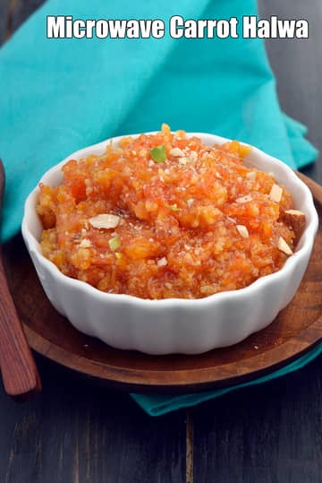 A white ceramic bowl filled with microwave carrot halwa, garnished with chopped nuts, placed on a wooden serving plate with a spoon on a dark table and a blue cloth in the background.
