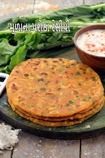 એક ઘેરા લાકડાના બોર્ડ પર સોનેરી-બદામી રંગના, શેકેલા ત્રણ મૂળાના પરાઠા ગોઠવેલા છે. પરાઠામાં લીલા ધાણા/ભાજી અને મૂળાના ટુકડા સ્પષ્ટ દેખાય છે. પાછળના ભાગમાં તાજી મૂળાની ભાજી અને એક લાકડાના બાઉલમાં સફેદ દહીં રાખેલું છે, જેના પર લાલ મરચું છાંટવામાં આવ્યું છે. ડાબી બાજુએ લાકડાની સપાટી પર તાજા ખમણેલા સફેદ મૂળાનો નાનો ઢગલો પણ જોવા મળે છે.