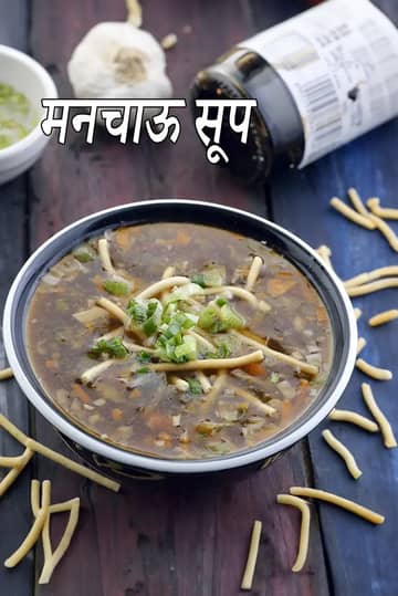 एक काले कटोरे में गरमा गरम वेजिटेबल मंचो सूप है, जिस पर कटी हुई हरी प्याज और क्रिस्पी तली हुई नूडल्स सजी हुई हैं। सूप में छोटे-छोटे कटी हुई सब्ज़ियाँ गाढ़े, गहरे रंग के शोरबे में हैं। कटोरे के चारों ओर तली हुई नूडल्स बिखरी हुई हैं, पीछे धुंधली पृष्ठभूमि में लहसुन का फूल और सोया सॉस की बोतल दिख रही है।