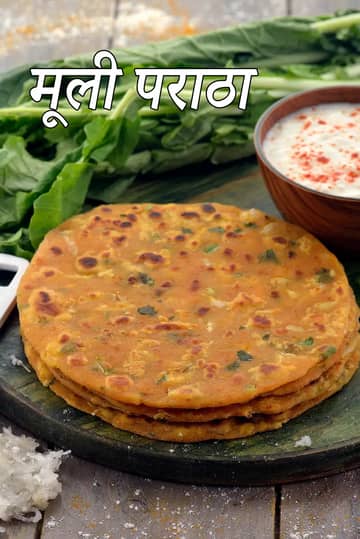 एक गहरे रंग के लकड़ीના बोर्ड पर तीन सुनहरे-भूरे, तवे पर सिँके हुए मूली के पराठे रखे हैं। पराठों में हरी जड़ी-बूटियाँ और मूली के टुकड़े साफ दिखाई दे रहे हैं। बैकग्राउंड में ताजी मूली की हरी पत्तियाँ और एक लकड़ी के कटोरे में सफेद गाढ़ा दही रखा है, जिस पर लाल मिर्च पाउडर छिड़का गया है। बाईं ओर, लकड़ी की सतह पर ताजी कसी हुई सफेद मूली का एक छोटा ढेर भी दिखाई दे रहा है।