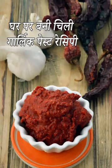 घर पर बने गहरे लाल मिर्च-लहसुन के पेस्ट का एक सुंदर दृश्य, जिसे एक छोटे सफेद नक्काशीदार कटोरे में रखा गया है। यह कटोरा एक गहरे रंग की लकड़ी की सतह पर रखा है, जिसके पास लहसुन की साबुत कलियां और सूखी लाल मिर्च बिखरी हुई हैं। पीछे की ओर एक बड़ा जार भी दिखाई दे रहा है जिसमें और पेस्ट भरा हुआ है, जो इसकी शुद्धता को दर्शाता है।