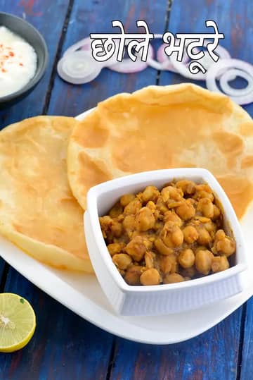 एक प्लेट में परोसा गया पारंपरिक छोले भटूरे, जिसमें फूले हुए सुनहरे तले हुए भटूरे और मसालेदार छोले की कटोरी है, साथ में प्याज़ के स्लाइस और नींबू, नीले लकड़ी के बैकग्राउंड पर।