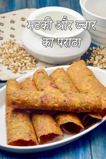 सफेद प्लेट में सलीके से रखी हुई मटकी और ज्वार की पराठियाँ, जो रोल के आकार में हैं। पराठियाँ सुनहरी-भूरी और हल्की कुरकुरी दिखाई देती हैं, जिनमें हरी जड़ी-बूटियाँ मिली हुई हैं। पीछे दही का कटोरा और अंकुरित मटकी रखी हुई है, जो नीले लकड़ी के बैकग्राउंड पर सजी है।