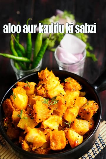 A bowl of aloo aur kaddu ki sabzi made with golden potato and pumpkin cubes cooked in Indian spices, garnished with fresh coriander, with green chilies and chopped onions placed in the background on a dark wooden surface.