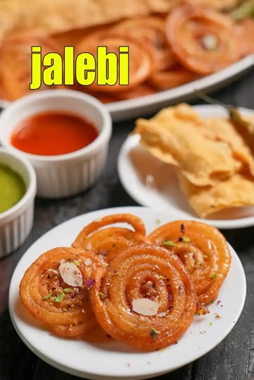 A close-up shot of crispy, golden-orange Jalebis served on a small white plate, garnished with sliced almonds, pistachios, and dried rose petals. In the background, there is a plate of Fafda and small bowls of red and green chutneys. The image captures a classic Indian breakfast combination, highlighting the syrupy texture of the sweet Jalebis.