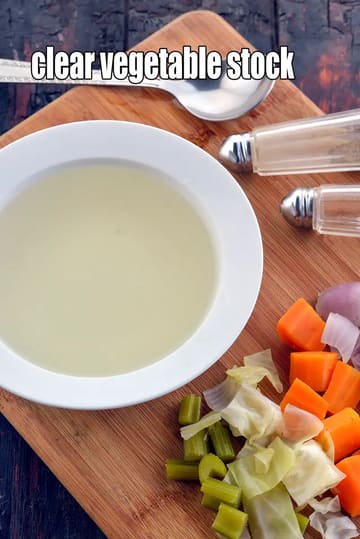 A bowl of clear vegetable stock placed on a wooden cutting board, surrounded by chopped vegetables like carrots, cabbage, celery, and onion, with a spoon and salt-and-pepper shakers beside it. The stock appears light, transparent, and freshly prepared.