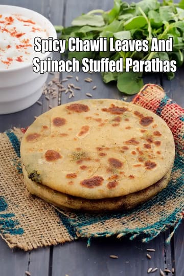 A stack of spicy chawli leaves and spinach stuffed parathas placed on a rustic cloth, with a bowl of white curd sprinkled with red chilli powder in the background, along with fresh green leafy vegetables and cumin seeds on a wooden surface.