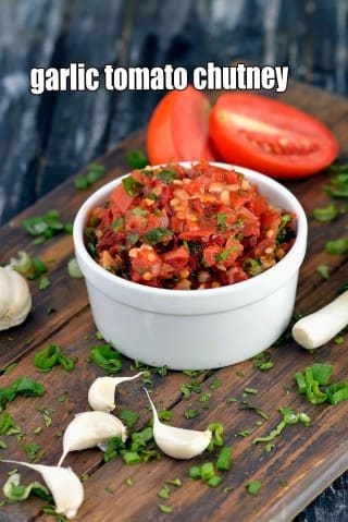 A close-up shot of Garlic Tomato Chutney served in a small white ceramic bowl, placed on a rustic wooden board. The chutney has a chunky, vibrant red texture with visible pieces of tomatoes, garlic, and green herbs. Fresh ingredients surround the bowl, including whole garlic cloves, sliced red tomatoes, and scattered chopped spring onions and coriander. The background features a dark, textured wooden surface that complements the rustic presentation.