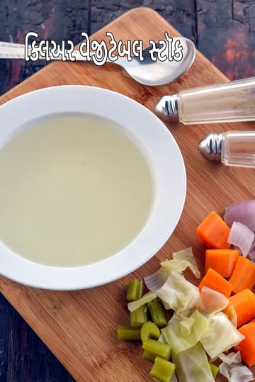 લાકડાના કટિંગ બોર્ડ પર મૂકાયેલું ક્લિયર વેજિટેબલ સ્ટોકનું બાઉલ, આસપાસ ગાજર, કોબી, સેલેરી અને ડુંગળી જેવા કાપેલા શાકભાજી રાખેલા છે. બાજુમાં ચમચી અને મીઠા-મરીની ડબ્બી દેખાય છે. સ્ટોક હળવું, સ્વચ્છ અને તાજું તૈયાર કરેલું લાગે છે.