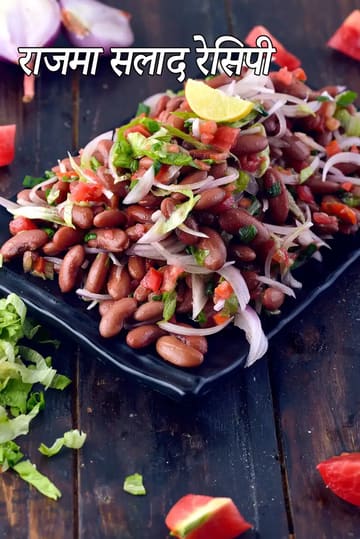उबले हुए राजमा से बना राजमा सलाद, जिसमें कटे हुए प्याज, टमाटर, लेट्यूस, हरी जड़ी-बूटियाँ और नींबू का टुकड़ा शामिल है, लकड़ी की पृष्ठभूमि पर काले बर्तन में परोसा गया।