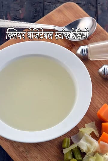 लकड़ी के कटिंग बोर्ड पर रखा साफ़ वेजिटेबल स्टॉक का एक कटोरा, जिसके पास कटे हुए गाजर, पत्ता गोभी, सेलेरी और प्याज़ रखे हैं। साथ में चम्मच और नमक-मिर्चदानी दिखाई दे रही है। स्टॉक हल्का, पारदर्शी और ताज़ा बना हुआ लग रहा है।