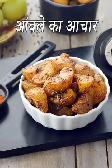 इस चित्र में एक सफेद नक्काशीदार कटोरे में रखे आंवले के अचार का क्लोज-अप दिखाया गया है, जो सौंफ और कलौंजी जैसे गहरे लाल-भूरे मसालों से लिपटा हुआ है। पृष्ठभूमि में, एक टोकरी में ताजे हरे आंवले और रसोई के काले बर्तन एक गहरी देहाती सतह पर रखे दिखाई दे रहे हैं।