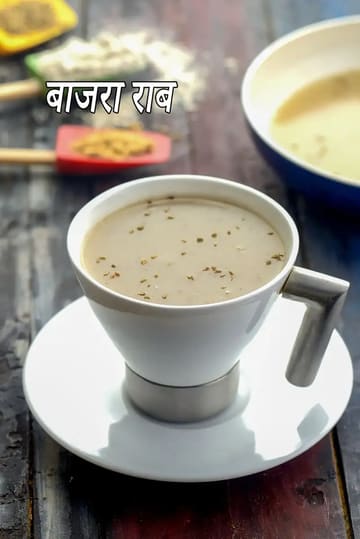 सफेद मिट्टी के कप में परोसी गई गरमा-गरम बाजरा राब, जो बाजरे के आटे से बना एक पारंपरिक स्वास्थ्यवर्धक पेय है। इसके ऊपर अजवाइन के दाने डाले गए हैं। कप एक सफेद तश्तरी  पर रखा है और बैकग्राउंड में बाजरे का आटा और एक लाल स्पैटुला दिखाई दे रहा है।