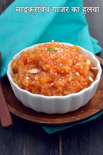 सफेद सिरेमिक कटोरे में माइक्रोवेव से बनी गाजर का हलवा, ऊपर से कटे हुए मेवों से सजाया हुआ, लकड़ी की प्लेट पर रखा है, साथ में चम्मच और पीछे नीले रंग का कपड़ा दिखाई दे रहा है।