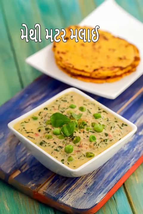 આ ચિત્રમાં ઉત્તર ભારતીય વાનગી 'મેથી મટર મલાઈ'નો ક્લોઝ-અપ બતાવવામાં આવ્યો છે. તેમાં એક ઘટ્ટ અને ક્રીમી ગ્રેવી છે, જેમાં ચમકતા લીલા વટાણા અને ઝીણી સમારેલી તાજી મેથીના પાન ઉમેરવામાં આવ્યા છે. વાનગીને તાજી ક્રીમથી સજાવવામાં આવી છે અને લાકડાના ટેબલ પર એક સુંદર સફેદ બાઉલમાં પીરસવામાં આવી છે.