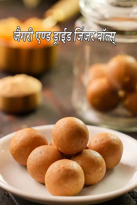 एक छोटी सफेद चीनी मिट्टी की प्लेट में करीने से रखे गए चिकने, गोल और सुनहरे भूरे रंग के गुड़ और सोंठ के लड्डू की तस्वीर। बैकग्राउंड में इन स्वास्थ्यवर्धक गोलियों से भरा एक कांच का जार और सोंठ पाउडर का एक धुंधला कटोरा दिखाई दे रहा है। हल्की रोशनी इन पारंपरिक इम्युनिटी बढ़ाने वाले लड्डुओं की चमक को दर्शाती है।