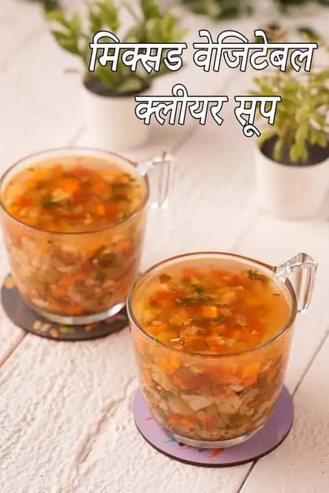 सफेद लकड़ी की टेबल पर कोस्टर पर रखे मिक्स वेजिटेबल क्लियर सूप के दो कांच के कप, पीछे छोटे पौधे और ऊपर लिखा टेक्स्ट “Mixed Vegetable Clear Soup”.