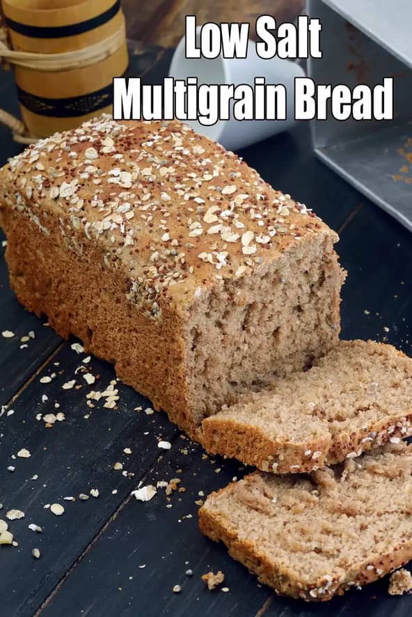 A rectangular loaf of low-salt multigrain bread sits on a dark, rustic wooden surface. The bread has a thick, hearty golden-brown crust topped with a generous layer of oats, seeds, and grains.