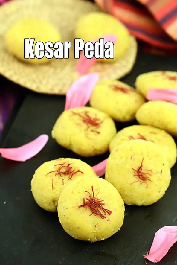 A close-up of yellow Kesar Peda sweets garnished with saffron strands, neatly arranged on a black serving slab, with pink rose petals scattered around and festive fabric in the background.