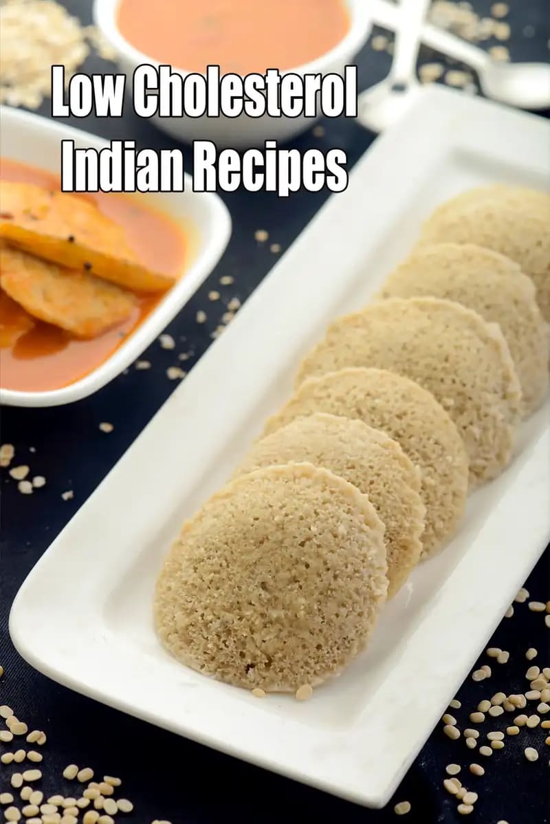A long white rectangular platter holds a row of soft, steamed oats idlis arranged neatly. In the background, there are two bowls: one containing a vibrant orange tomato-based chutney and another with a different flavorful accompaniment. The setting is on a dark surface, with some raw lentils or grains scattered around to highlight the healthy ingredients. Large white text at the top of the image reads, Low Cholesterol Indian Recipes.