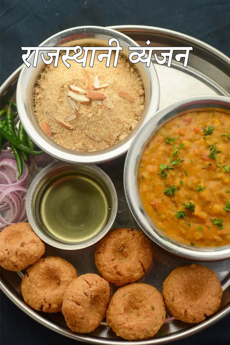 पारंपरिक स्टील की थाली में परोसा गया राजस्थानी भोजन, जिसमें दाल, बादाम से सजा चूरमा, छोटी कटोरी में घी और बाटी शामिल हैं, साथ में कटे हुए प्याज़ और हरी मिर्च, तथा तस्वीर पर राजस्थानी भोजन लिखा हुआ दिखाई देता है।