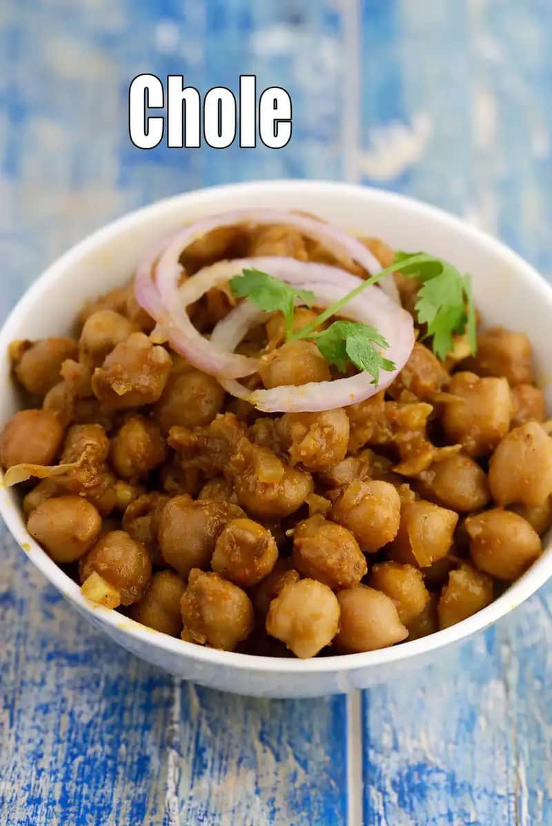 A white bowl filled with dry-style Punjabi chole, made from cooked chickpeas tossed in spices, garnished with onion rings and fresh coriander, placed on a rustic blue wooden surface.