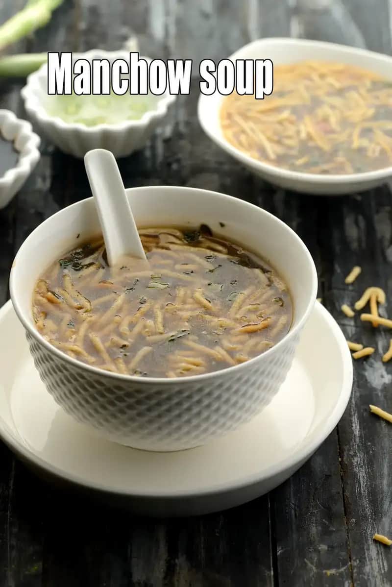 A high-angle shot of a steaming bowl of Veg Manchow Soup served in a white textured ceramic bowl with a matching spoon. The dark, brownish broth is filled with finely chopped vegetables and topped with a generous portion of crispy fried noodles. In the background, another bowl of soup and small condiment bowls containing chillies in vinegar and soy sauce are visible on a rustic dark wooden table.