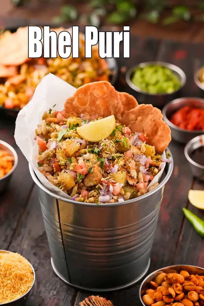 A vibrant serving of Bhel Puri presented in a small metal bucket lined with white paper, topped with a lemon wedge and crispy papdis, surrounded by small bowls of chutneys and sev.