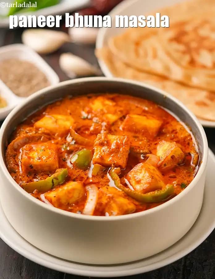 An image featuring a white bowl filled with Paneer Bhuna Masala, a rich and flavorful Indian gravy dish. The curry has a thick, orange-red base containing cubes of succulent paneer, sliced onions, and green bell peppers. In the background, a plate of soft parathas is visible, suggesting a complete meal. The dish is presented on a dark surface, with ingredients like garlic cloves and spices blurred in the distance.