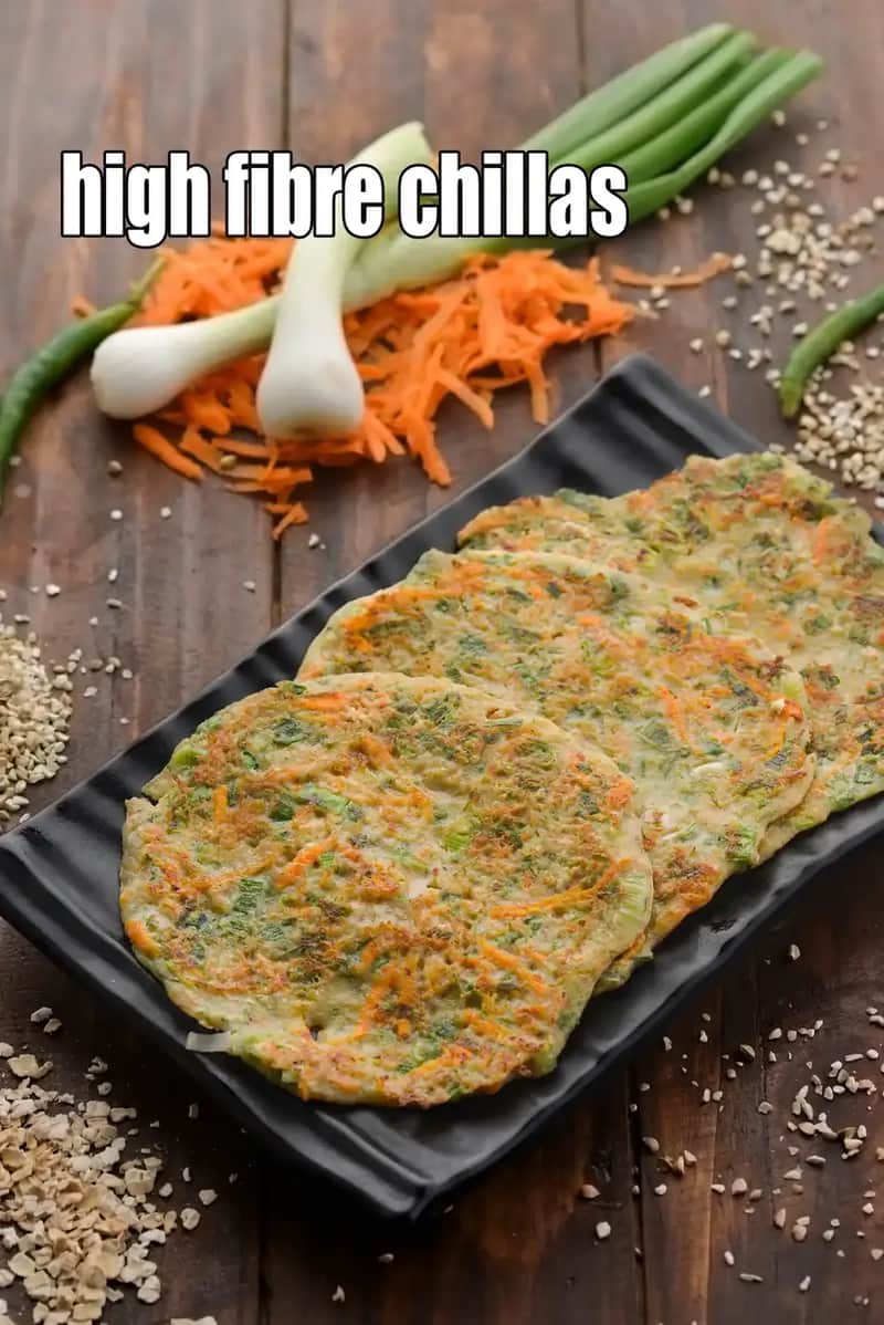 A black rectangular plate holds a stack of four savory high-fiber chillas (Indian pancakes) arranged diagonally. The chillas are golden-brown and studded with visible shredded carrots and green herbs. In the background, fresh ingredients like spring onions, grated carrots, and green chillies are scattered on a rustic wooden surface. Grains and seeds are also sprinkled around the plate, emphasizing the healthy, fibrous nature of the dish.