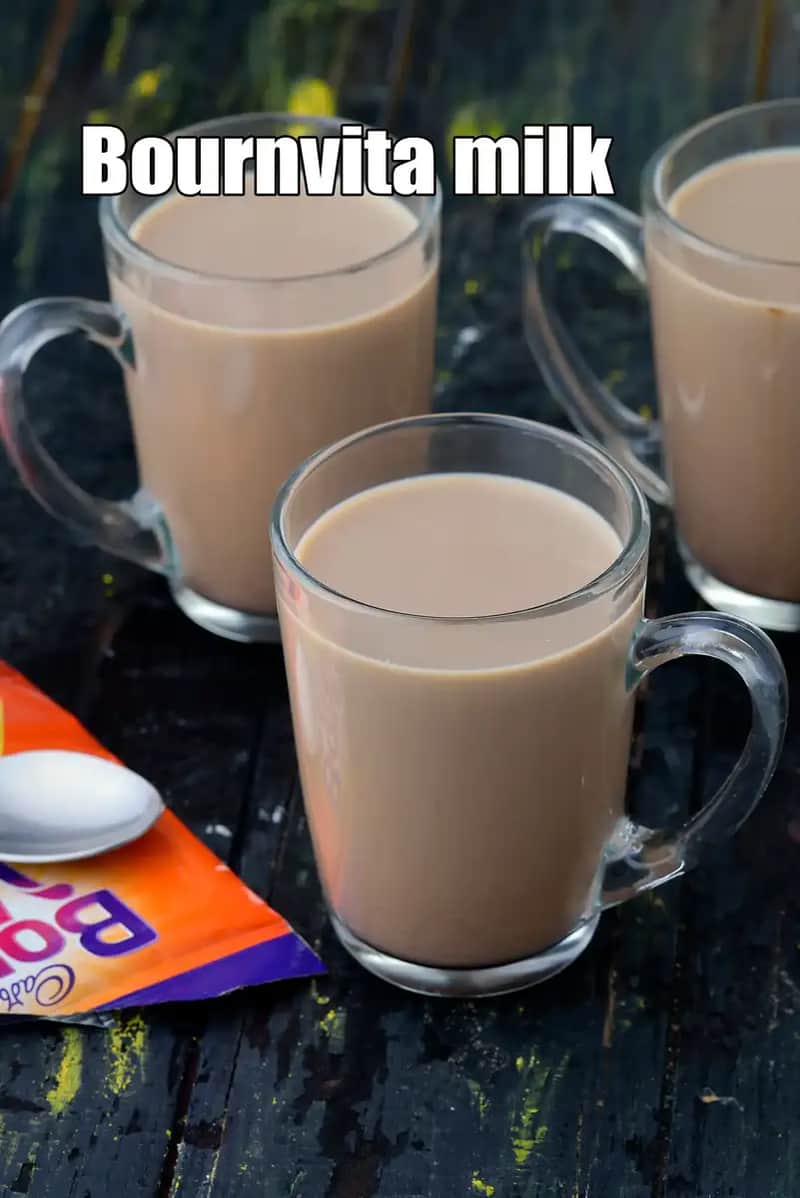 A high-angle shot featuring three glass mugs filled with warm, brown Bournvita milk, arranged on a dark, rustic wooden surface. In the foreground, an open orange and purple Cadbury Bournvita sachet lies next to a small white spoon.