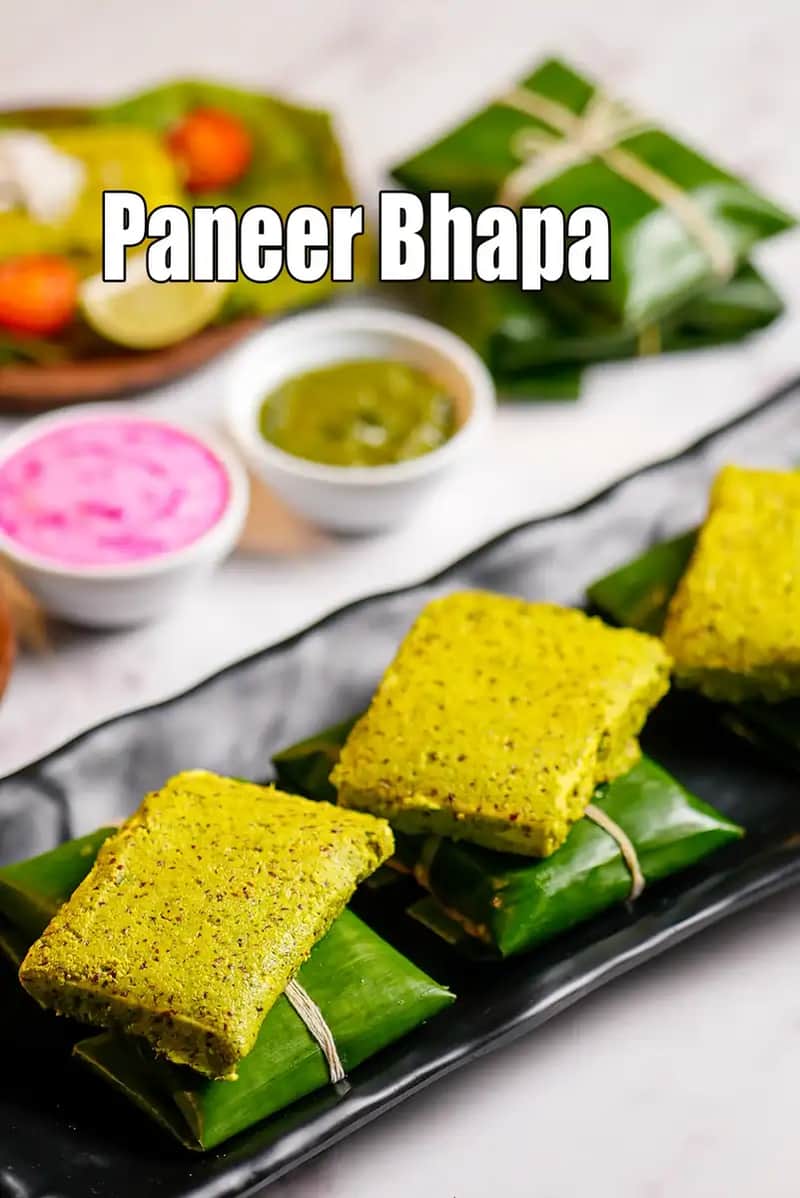 Paneer Bhapa served on a black platter, showing steamed paneer pieces wrapped in banana leaves, coated with green mustard paste, accompanied by green chutney and pickled onions.