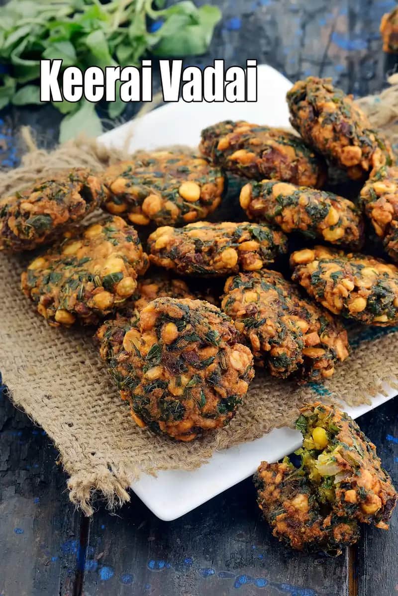 A close-up view of crispy Keerai Vadai (amaranth leaves fritters) stacked on a white plate lined with a rustic cloth. The deep-fried vadas are golden-brown with visible lentils and chopped greens, placed on a dark wooden background, with fresh green leaves seen in the background.